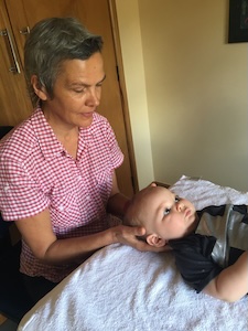 Practitioner treating a baby, her hands are under his head while he lies on his back on a treatment table looking up.